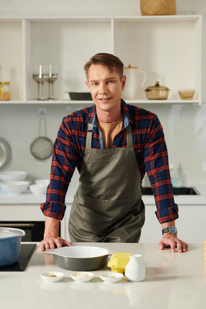 Hymyilevä mies seisoo keittiön tiskillä herkullisen kastikkeen ainesosalla pienissä kulhoissa hänen edessään. - Valokuva, kuva