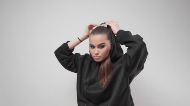 A young woman fashion model with bold makeup, winged eyeliner and a stylish ponytail hairstyle takes off the hood of a fashionable black hoodie and poses in the studio on a white background. - Footage, Video