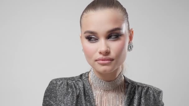 A beautiful young woman with perfect smooth skin, with bright evening makeup with winged eyeliner and spectacular jewelry, posing on a white background, glance looking at the camera. - Footage, Video