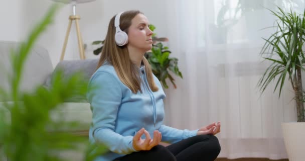 Una joven feliz con auriculares practicando yoga y meditación en casa, sentada en el suelo de la sala de estar en pose de loto y relajada con los ojos cerrados. El concepto de meditación consciente y bienestar - Imágenes, Vídeo