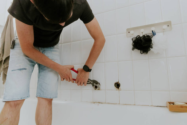 Один молодой кавказский парень с брюнеткой откручивает трубу от крана регулируемым гаечным ключом в белой черепичной стене в ванной комнате дома, крупным планом.. - Фото, изображение