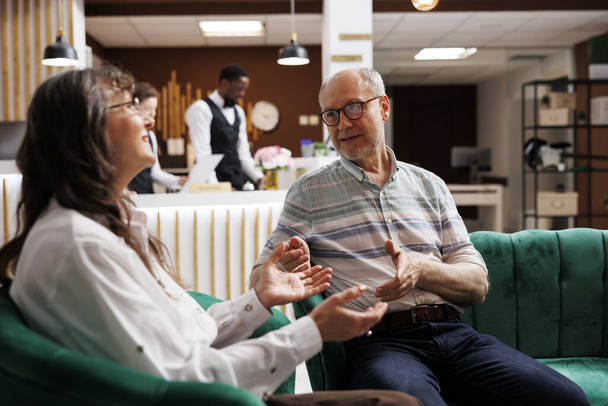 Ein älteres kaukasisches Paar diskutiert in der Lounge eines Luxus-Resorts über seine Reisepläne. Rentnerehepaar wartet an der Hotelrezeption auf den Check-in und plant eine wunderbare Hochzeitsreise. - Foto, Bild