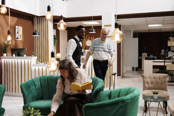 Vriendelijk Afro-Amerikaanse piccolo assisteert senior man in hotellobby met bagage naar de gereserveerde kamer. Oudere vrouwelijke bezoeker leest tijdens het wachten op accommodatie in gezellige lounge met fauteuils. - Foto, afbeelding