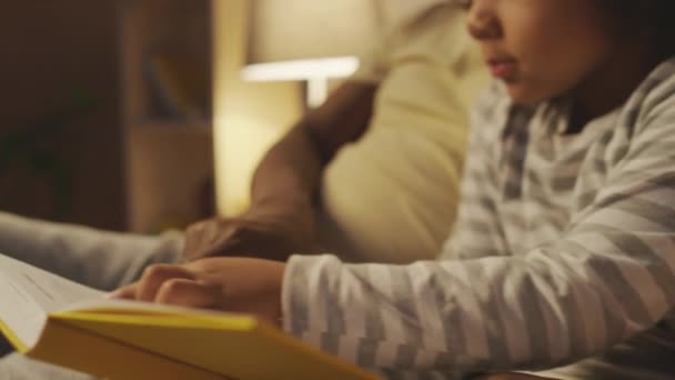 Boczne pochylenie zbliżenie małego kręconego Afroamerykanina chłopca w piżamie siedzi z książki w sypialni i słucha bajki, a następnie omawianie fabuły z tatą przed snem - Materiał filmowy, wideo