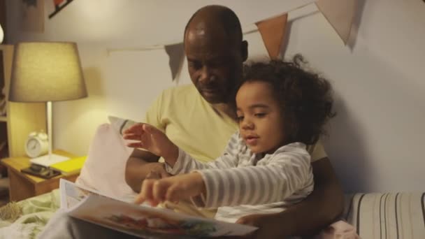 Mediana toma de niño rizado poco excitado en pijamas señalando y hablando de cómics con dinosaurios acostados en la cama con el padre afroamericano en el dormitorio decorado antes de dormir - Metraje, vídeo