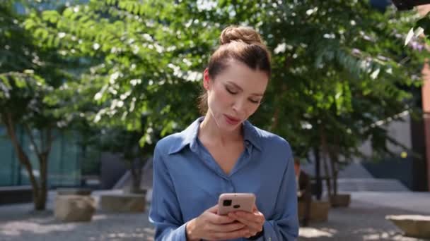Beztroski menedżer smsy z telefonu komórkowego siedzi słoneczny rynek zbliżenie. Atrakcyjna młoda kobieta patrząca na ekran smartfona z uśmiechem na zewnątrz. Happy businesswoman czytanie wiadomości relaksujący park - Materiał filmowy, wideo