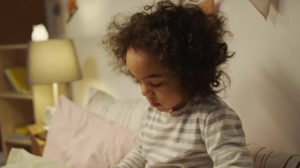Бічний нахил п'ятирічного афроамериканського хлопчика в піжамі сидить у ліжку перед сном і дивиться на мультфільми динозаврів, які посилаються через комікс перед сном - Кадри, відео