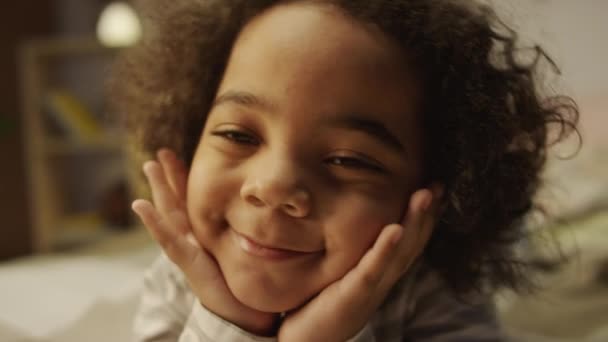 Primo piano ritratto di felice attivo afroamericano dai capelli ricci ragazzo in pigiama appoggiando la testa con le mani e poi ridere e sorridere mentre sdraiato sullo stomaco in camera da letto - Filmati, video