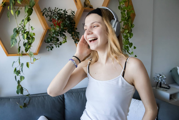 Jonge vrouw, blond, blank, luisteren naar muziek met koptelefoon in haar appartement, met een aantal planten en de bank op de achtergrond. - Foto, afbeelding