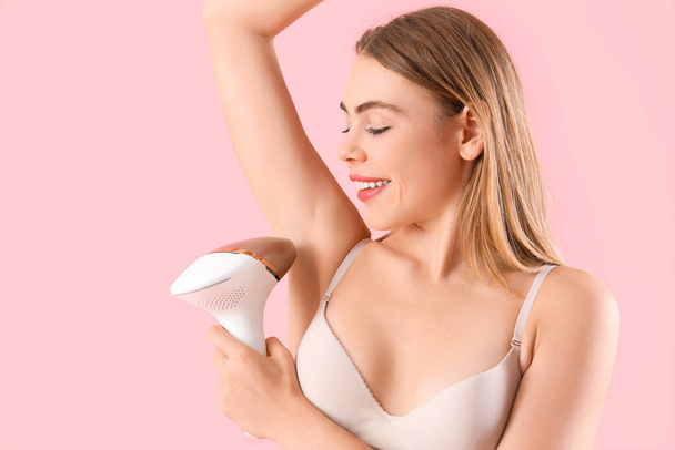 Beautiful young woman epilating her armpits with modern photoepilator on pink background - Photo, Image