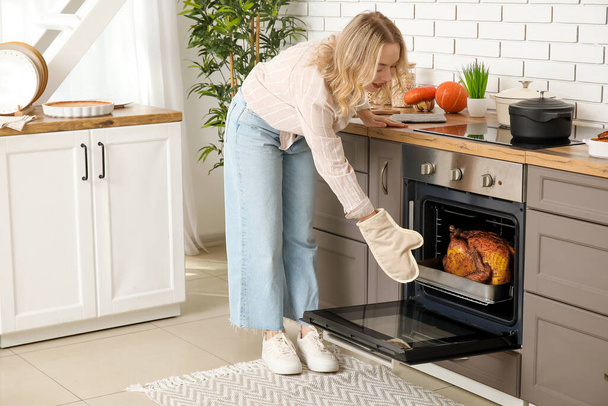 Beautiful young woman taking tasty baked turkey from oven in kitchen. Thanksgiving Day celebration - Photo, Image