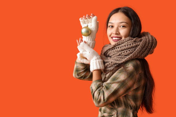 Mooie jonge Aziatische vrouw in warme sjaal met kerstbal op oranje achtergrond - Foto, afbeelding