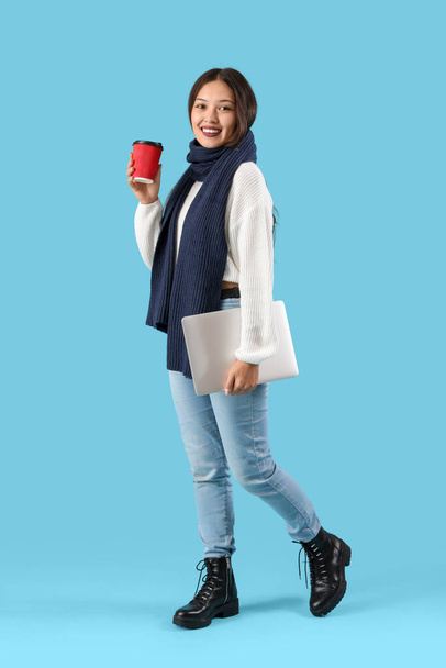 Kaunis nuori aasialainen nainen lämpimässä huivissa kupin kahvia ja kannettavaa sinisellä pohjalla - Valokuva, kuva