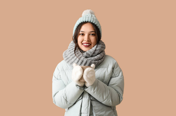 Mooie jonge Aziatische vrouw in warme sjaal met kopje thee op bruine achtergrond - Foto, afbeelding