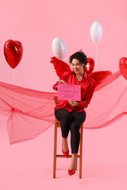 ピンクの背景のバレンタインデーのためのキューピッドの翼とカードを持つ若いアフリカ系アメリカ人女性 - 写真・画像