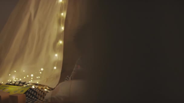 Panoramique des images moyennes de l'enfant afro-américain de 5 ans avec des cheveux bouclés portant un pyjama couché dans un tipi à la main et jouant sur une tablette dans une chambre confortable décorée de lumières à ficelle la nuit - Séquence, vidéo
