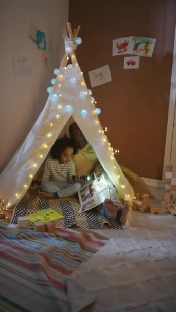 Yatma vakti yatak odasında el yapımı çadırı içinde çizgi roman okuyan babasıyla el feneri olan Afrikalı Amerikalı çocuğun dikey görüntüsü. - Video, Çekim