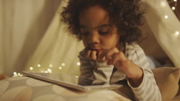 Портативный крупный план афроамериканского маленького кудрявого ребенка в пижаме, выбирающего видео или мультфильм на планшете, лежащем на подушках ночью в мечтательной спальне, украшенной гирляндой - Кадры, видео