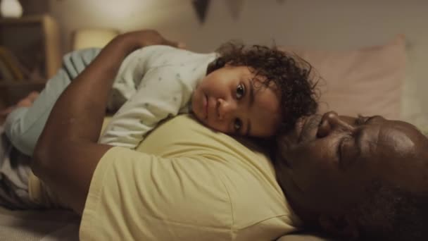 Retrato de mano lateral de la pequeña niña Biracial mirando a la cámara y acostada en el vientre de los padres afroamericanos que arrullan a su hijo para dormir por la noche - Imágenes, Vídeo