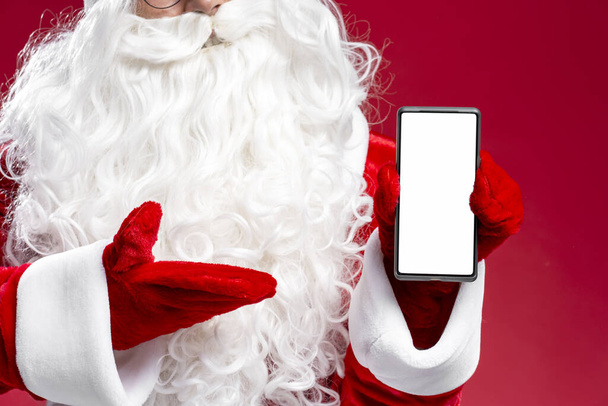Lähikuva Santa käsi tilalla matkapuhelin näyttää näytön eristetty punaisella taustalla, mockup. Verkkokaupoissa, myynti, mobiilisovellus käsite  - Valokuva, kuva