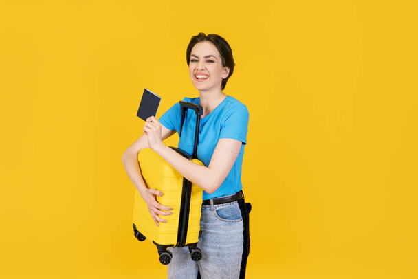 radosna kobieta w niebieskiej koszulce z żółtą walizką i paszportem na żółtym tle. Podróże cieszyć koncepcja - Zdjęcie, obraz