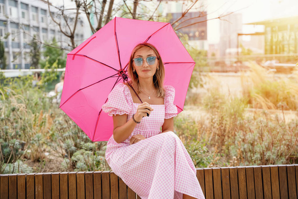 Asijka v růžových šatech a slunečních brýlích s růžovým deštníkem sedí v centru města. nečinný koncept životního stylu - Fotografie, Obrázek