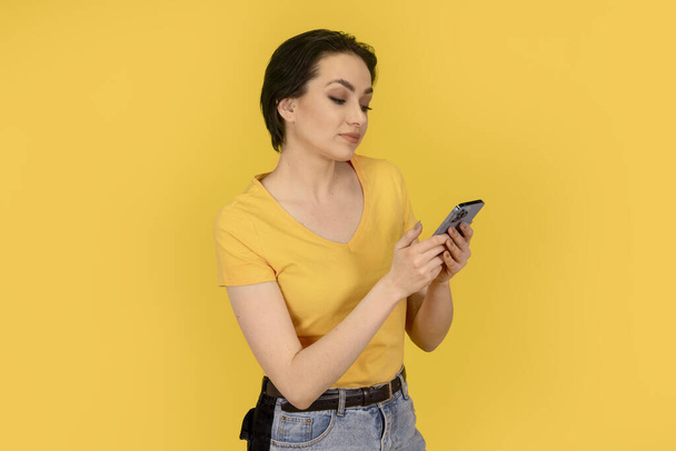 überraschte Frau in gelbem T-Shirt und Jeans mit Handy auf gelbem Hintergrund - Foto, Bild