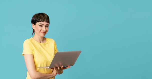 Smiling woman in a yellow t-shirt using laptop, pc computer on blue background with copy space. Work studying, virtual training, e-learning, watching online education webinar concept - Photo, Image