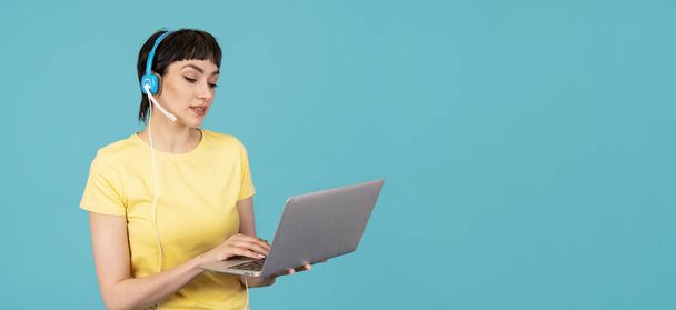 イエローのTシャツとヘッドフォンの素晴らしい女性は,コピースペースを持つ青い背景のPCコンピュータを使用しています. ワークスタディ,バーチャルトレーニング,eラーニング,オンライン教育ウェビナーのコンセプトを見る - 写真・画像