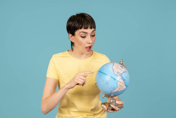 Kobieta w żółtym t-shircie wskazuje palcem na kulę ziemską wybierając miejsce wypoczynku odizolowane na niebieskim tle Pasażer podróżujący za granicę. co wybierzesz. podróż, koncepcja edukacji - Zdjęcie, obraz