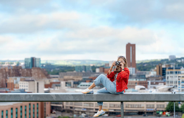 glückliche Asiatin in roter Jacke sitzt vor der Stadt Sheffield und macht Fotos an einem tollen Sommertag - Foto, Bild