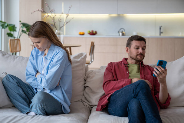 Eifersüchtige Ehefrau traurig über Nachrichten ihres Mannes, der zu Hause auf der Couch sitzt. Ehepartner ignorieren einander. Empörte Frauen entpuppen sich als Männer, die aufs Smartphone schauen. Verrat Untreue Misstrauen Konzept. - Foto, Bild