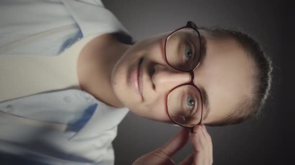 Vertikální střední detailní portrét mladé bělošky dívající se do kamery při úpravě brýlí, stojící na šedém pozadí studia - Záběry, video
