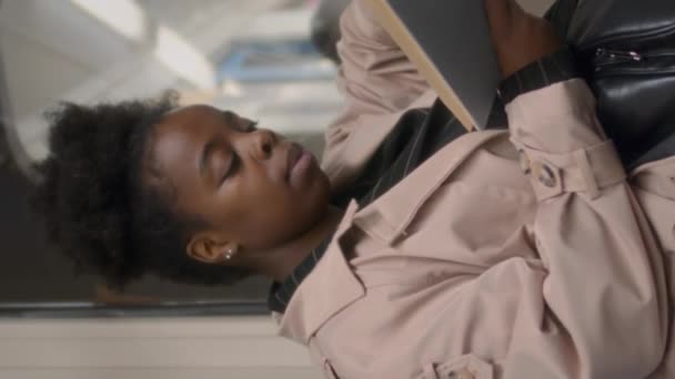 Vertical médio tiro do livro de leitura jovem mulher negra enquanto sentado sozinho pela janela no trem do metrô - Filmagem, Vídeo