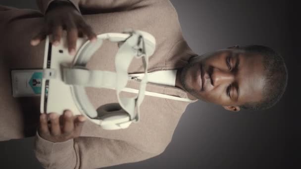 Вертикальный средний портрет молодого чернокожего человека, позирующего перед камерой на сером фоне студии с белой VR гарнитурой в руках - Кадры, видео