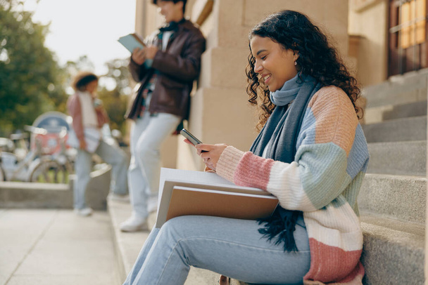 Giovane studentessa in cerca di telefono seduto vicino al campus universitario su sfondo amici - Foto, immagini