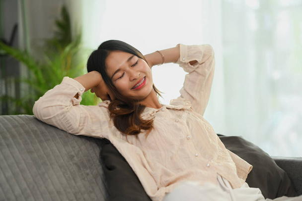 Glückliche junge Frau mit den Händen hinter dem Kopf entspannt sich zu Hause auf der Couch - Foto, Bild