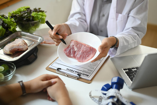 Καλλιεργημένη λήψη διατροφολόγου που κρατάει πιάτο με ωμό κρέας δίνοντας συμβουλές στον ασθενή στην κλινική - Φωτογραφία, εικόνα