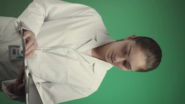 Κάθετη μέτρια αργό πορτρέτο της νεαρής καυκάσιας γιατρού φορώντας λευκή ρόμπα εργαστηρίου και σήμα ταυτότητας στο λαιμό της ποζάροντας για κάμερα με ιατρική κάρτα ασθενούς σε πράσινο φόντο - Πλάνα, βίντεο