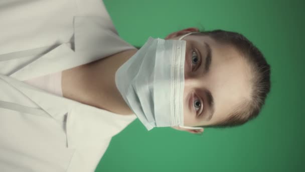 Pystysuora rinta ylös hidas muotokuva nuori valkoihoinen naislääkäri katselee kameraa ottaessaan pois kasvonaamio, seisoo vihreä studio tausta - Materiaali, video