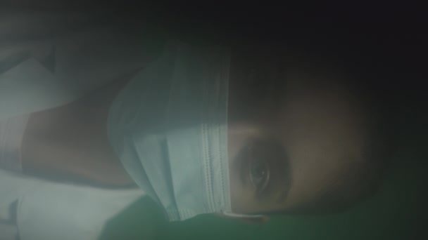 Mavi gözlü beyaz kadın doktorun dikey portresi. Yüz maskesi takmış hastalara bakıyor. Röntgen ve kamera, açık yeşil arka planda duruyor. - Video, Çekim