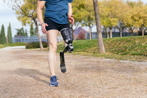 Unrecognizable male runner with prosthetic sports leg on park path. Incognito athletic amputee enjoys running, embodying adaptive sports spirit. - Photo, Image