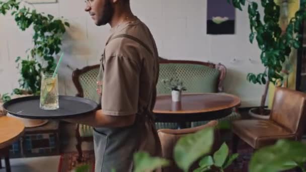 Średniej jakości zdjęcie męskiego afroamerykańskiego kelnera w fartuchu, okularach i bandanie w pracy w restauracji lub kawiarni, niosącego napój w szklance do klientki, kładącego na stół i wychodzącego - Materiał filmowy, wideo