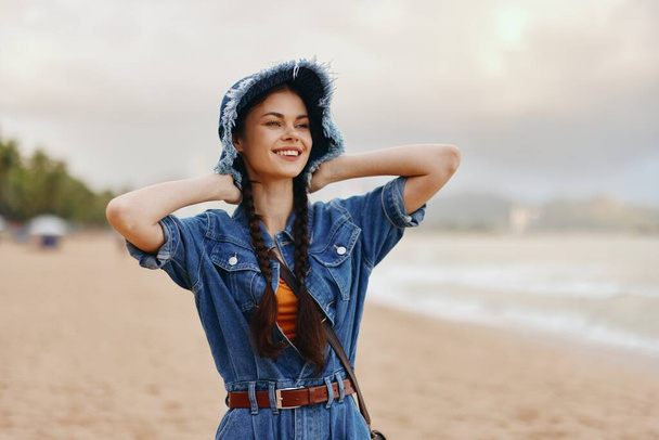 Stylische Kaukasierin mit attraktiven Haaren, die den Sommerurlaub am Strand genießt, glücklich und entspannt aussieht, mit hübschem Gesicht und modischem Hut, vor einem wunderschönen Hintergrund in der Natur - Foto, Bild