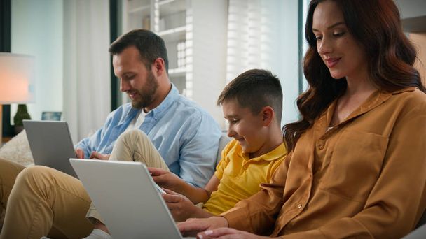 Addicted to modern technology dad mom son using different gadgets happy Caucasian family spend time together parents working remote on laptop at home on couch kid boy child playing video game on phone - Photo, Image
