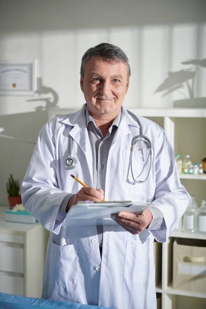 Portrait of positive mature veterinarian filling medical form on clipboard - Photo, Image