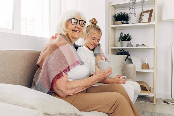 女性は,おばあちゃんの幸せな孫娘のマグカップを抱擁する愛のソファを置きました 老人ホーム 家族 冬のソファー - 写真・画像