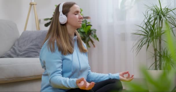 Μια χαρούμενη νεαρή γυναίκα που φοράει ακουστικά και κάνει γιόγκα και διαλογισμό στο σπίτι, κάθεται στο πάτωμα του σαλονιού στο λωτό και χαλαρώνει με τα μάτια κλειστά. Η έννοια του προσεκτικού διαλογισμού και της ευημερίας - Πλάνα, βίντεο