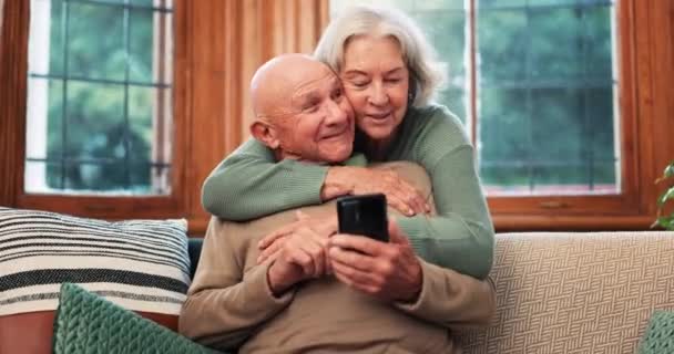 Idős pár, ölelés és pihenés kanapén telefon, alkalmazás vagy streaming egy videót a közösségi média internet. Olvasás, mobiltelefon és boldog öregek nyugdíjas szeretettel vagy görgetés hírek a nappaliban. - Felvétel, videó