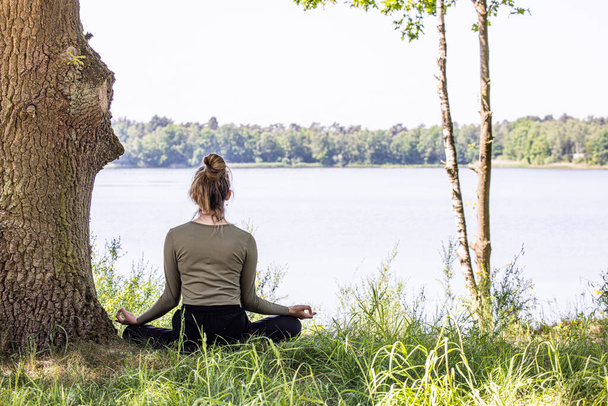 A young woman finds inner peace as she practices yoga in the heart of nature. Sitting gracefully on the lush green grass beside a majestic tree and a tranquil forest lake, she engages in a meditation - Photo, Image